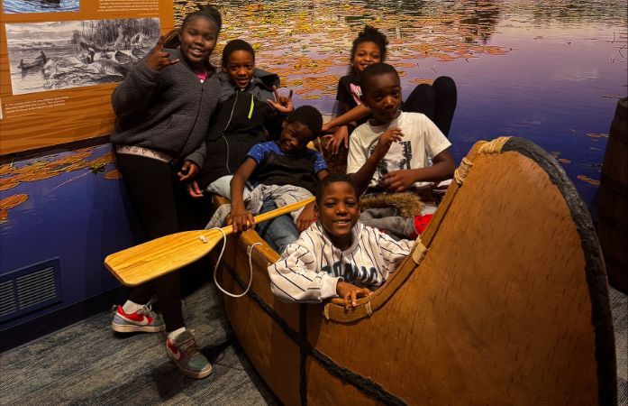  6 students in model canoe at museum smiling at camera